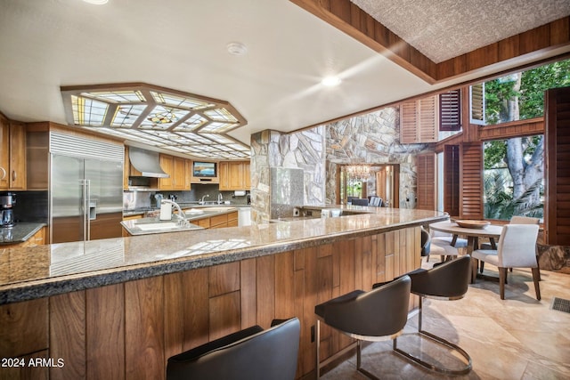 kitchen featuring kitchen peninsula, a kitchen breakfast bar, tasteful backsplash, wall chimney range hood, and built in fridge