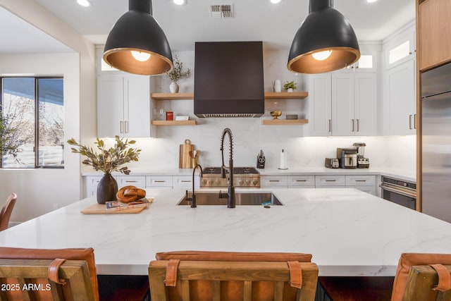 kitchen featuring decorative light fixtures, white cabinetry, light stone counters, stainless steel appliances, and a center island with sink