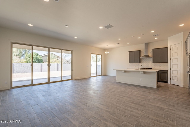 kitchen featuring backsplash, hardwood / wood-style flooring, a notable chandelier, a center island with sink, and wall chimney exhaust hood