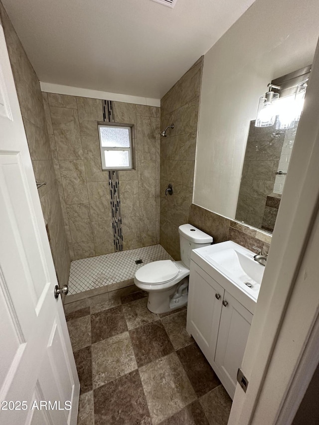 full bathroom with a tile shower, vanity, toilet, stone finish floors, and tile walls