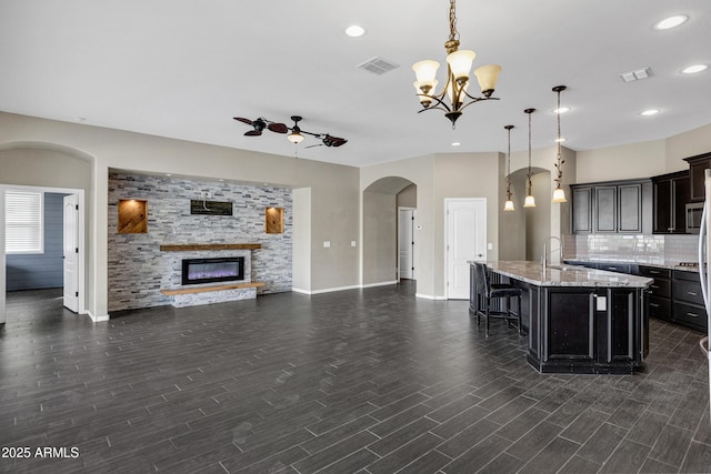 kitchen featuring decorative light fixtures, an island with sink, a fireplace, ceiling fan with notable chandelier, and backsplash