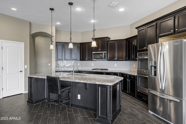 kitchen featuring sink, hanging light fixtures, stainless steel appliances, light stone countertops, and a center island with sink