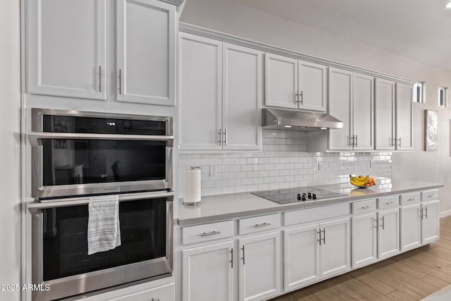 kitchen with decorative backsplash, black electric cooktop, stainless steel double oven, light countertops, and under cabinet range hood