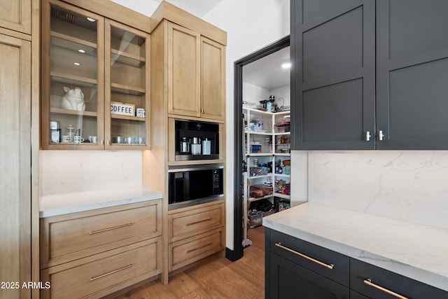 kitchen featuring glass insert cabinets, light stone counters, dark cabinetry, light brown cabinetry, and light wood-style floors