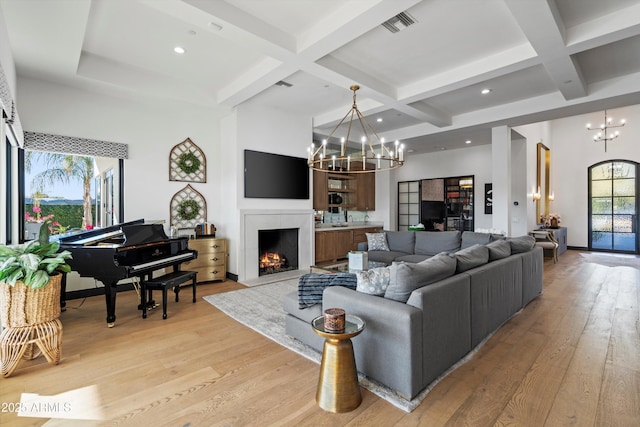 living room featuring a warm lit fireplace, light wood-style floors, coffered ceiling, and a notable chandelier