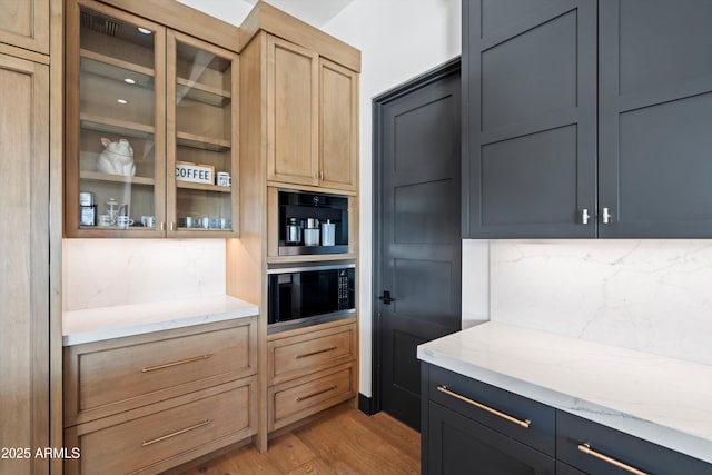 kitchen featuring glass insert cabinets, dark cabinets, light stone countertops, light wood-type flooring, and backsplash