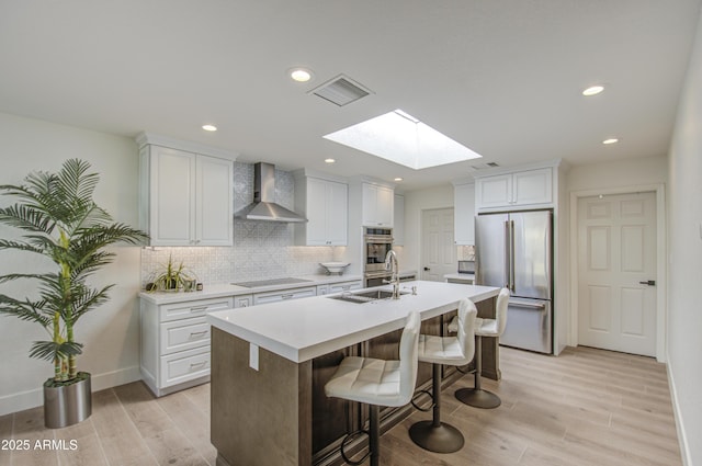 kitchen featuring a breakfast bar, an island with sink, white cabinets, stainless steel appliances, and wall chimney range hood