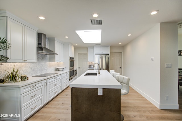 kitchen with a skylight, sink, white cabinets, stainless steel appliances, and wall chimney range hood