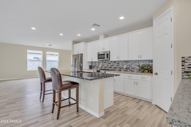 kitchen featuring a kitchen island with sink, dark stone counters, light wood-type flooring, stainless steel appliances, and white cabinets