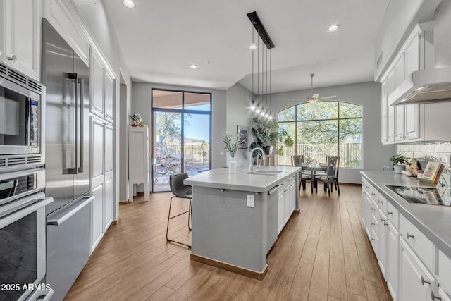 kitchen featuring sink, a kitchen island with sink, stainless steel appliances, decorative light fixtures, and wall chimney exhaust hood