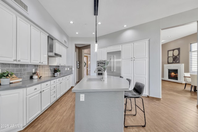 kitchen featuring wall chimney range hood, sink, hanging light fixtures, stainless steel built in refrigerator, and a center island with sink