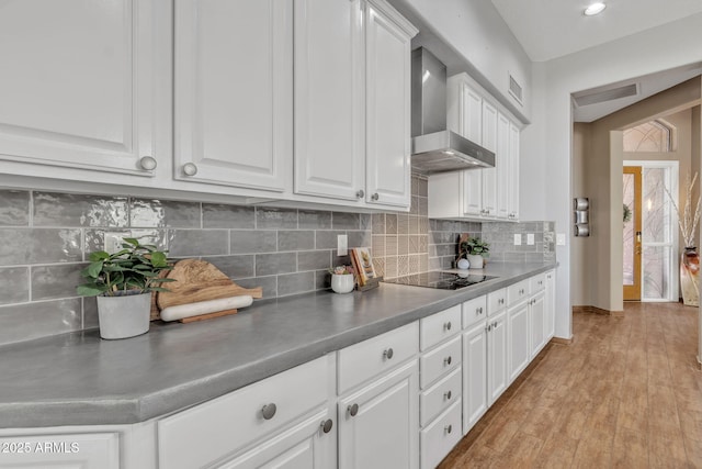 kitchen featuring wall chimney exhaust hood, black electric cooktop, white cabinets, light hardwood / wood-style floors, and backsplash