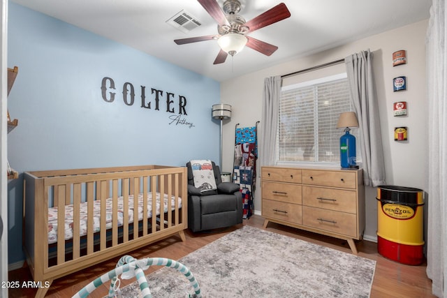 bedroom featuring a nursery area, ceiling fan, visible vents, and wood finished floors