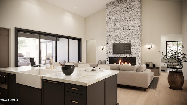 kitchen with a kitchen island with sink, high vaulted ceiling, a stone fireplace, sink, and light hardwood / wood-style flooring