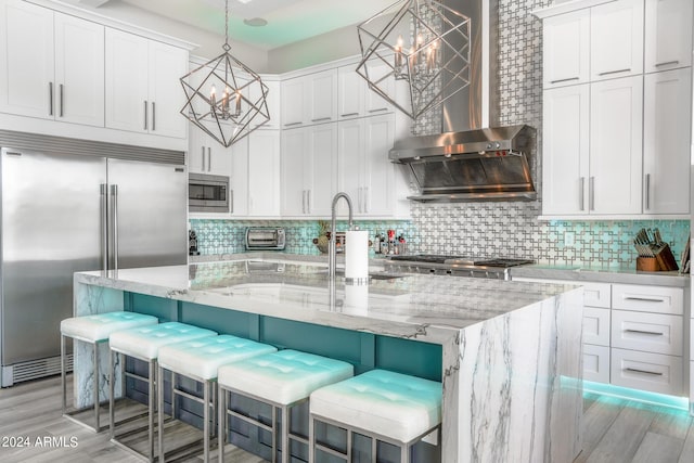 kitchen featuring built in appliances, decorative light fixtures, an island with sink, and white cabinets