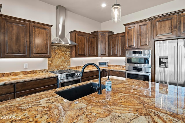 kitchen with light stone counters, pendant lighting, sink, wall chimney exhaust hood, and stainless steel appliances