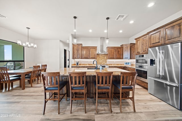 kitchen with wall chimney range hood, a kitchen island with sink, pendant lighting, light stone countertops, and appliances with stainless steel finishes