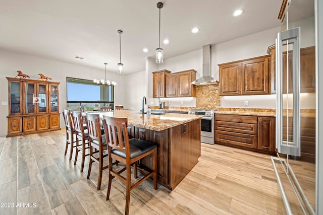 kitchen with wall chimney range hood, light wood-type flooring, stainless steel range with electric cooktop, sink, and an island with sink