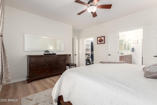 bedroom with baseboards, ensuite bath, a walk in closet, light wood-type flooring, and a ceiling fan