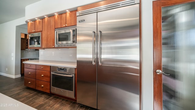 kitchen featuring brown cabinets, dark wood finished floors, light countertops, backsplash, and built in appliances