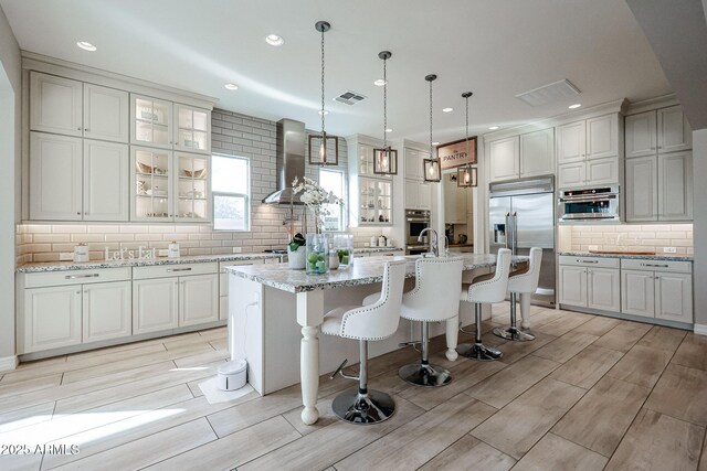 kitchen with visible vents, stainless steel appliances, wall chimney exhaust hood, an island with sink, and backsplash