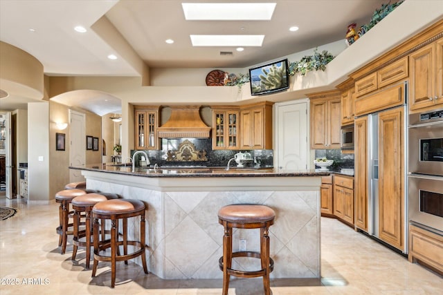 kitchen with built in appliances, a kitchen island with sink, a kitchen breakfast bar, backsplash, and custom range hood