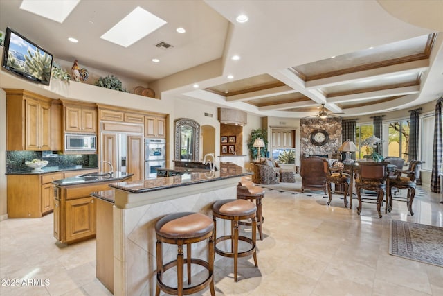 kitchen with a skylight, a sink, built in appliances, coffered ceiling, and a large island with sink