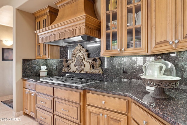 kitchen featuring glass insert cabinets, dark stone countertops, premium range hood, stainless steel gas stovetop, and backsplash