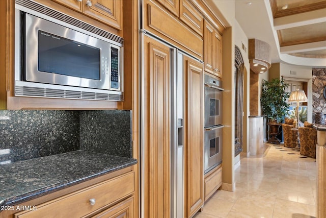 kitchen featuring stainless steel appliances, brown cabinetry, ornamental molding, light tile patterned flooring, and dark stone countertops