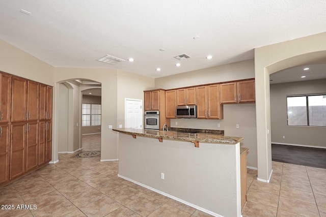 kitchen featuring visible vents, brown cabinetry, arched walkways, stainless steel microwave, and light stone counters