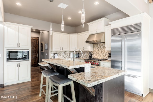 kitchen with hanging light fixtures, exhaust hood, dark hardwood / wood-style flooring, light stone countertops, and built in appliances