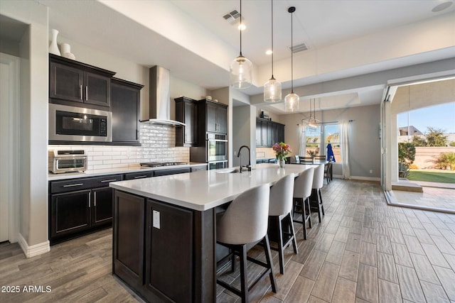 kitchen with built in microwave, wall chimney exhaust hood, stainless steel gas cooktop, an island with sink, and pendant lighting