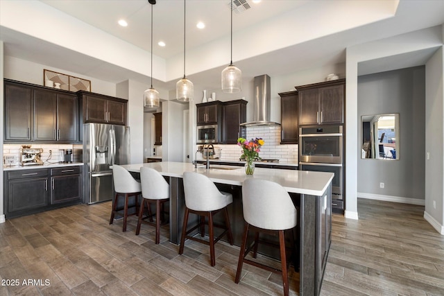 kitchen featuring a kitchen breakfast bar, pendant lighting, stainless steel appliances, a kitchen island with sink, and wall chimney range hood