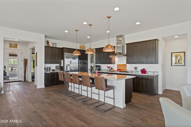 kitchen featuring appliances with stainless steel finishes, dark brown cabinetry, a kitchen island with sink, wall chimney range hood, and pendant lighting