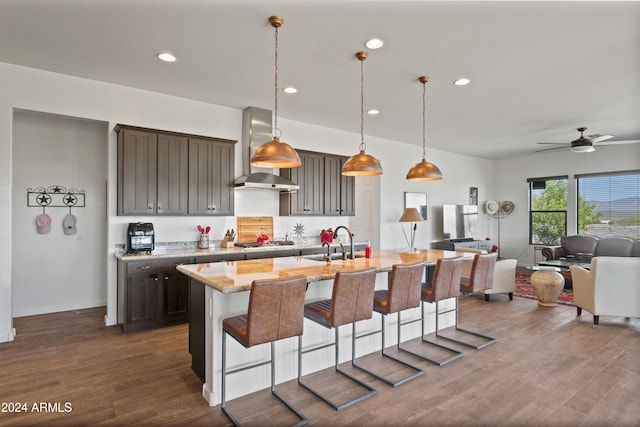 kitchen featuring wall chimney range hood, light stone counters, stainless steel gas stovetop, a kitchen island with sink, and a breakfast bar