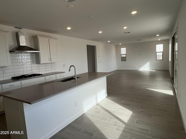 kitchen with a kitchen island with sink, a sink, gas stovetop, white cabinets, and wall chimney exhaust hood