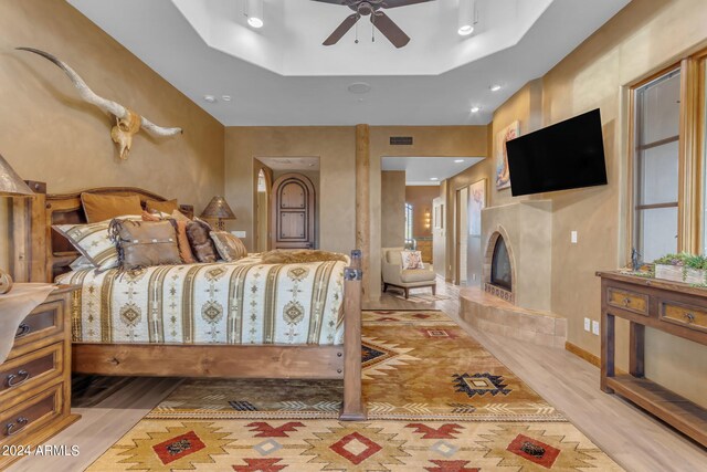 bedroom with light wood-type flooring, a tiled fireplace, a tray ceiling, and ceiling fan