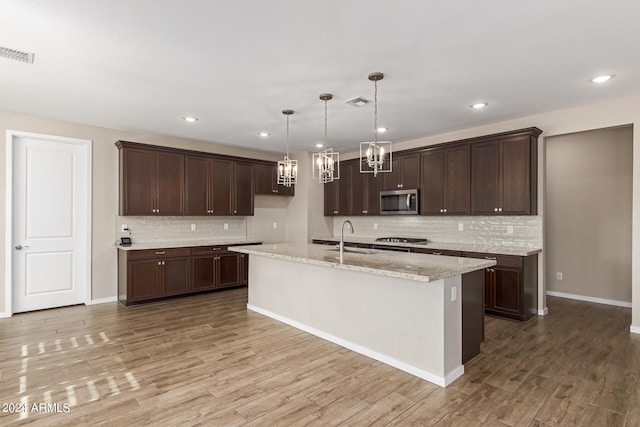 kitchen with light wood-type flooring, stainless steel appliances, decorative light fixtures, light stone counters, and a kitchen island with sink