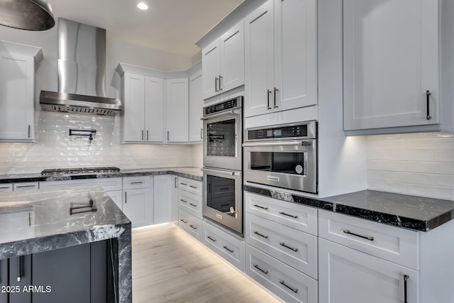 kitchen with light wood-style flooring, white cabinets, wall chimney range hood, appliances with stainless steel finishes, and dark stone counters