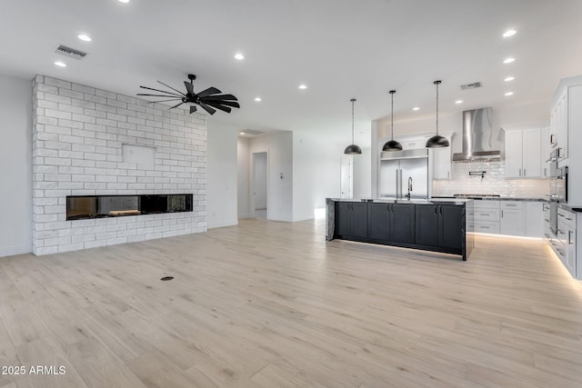 kitchen featuring white cabinets, wall chimney exhaust hood, open floor plan, a kitchen island with sink, and pendant lighting