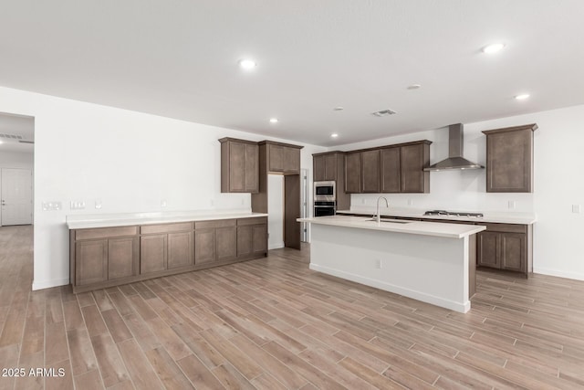 kitchen with sink, a center island with sink, light hardwood / wood-style flooring, stainless steel appliances, and wall chimney range hood