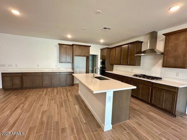 kitchen with sink, dark brown cabinets, an island with sink, stainless steel gas cooktop, and wall chimney exhaust hood