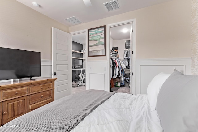 bedroom with wainscoting, a walk in closet, a closet, ceiling fan, and a decorative wall