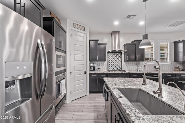 kitchen with appliances with stainless steel finishes, a sink, wall chimney range hood, backsplash, and light stone countertops