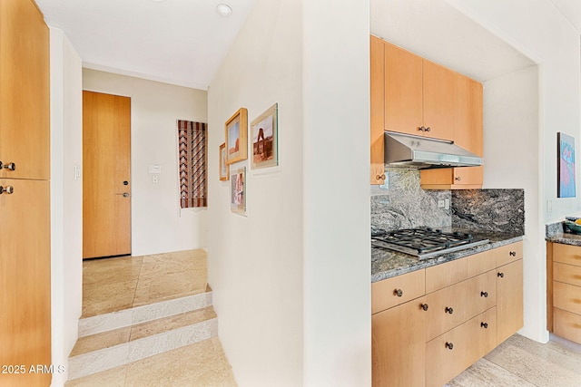 kitchen with under cabinet range hood, stainless steel gas cooktop, decorative backsplash, dark stone counters, and light brown cabinetry