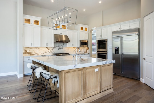 kitchen with built in appliances, white cabinets, light stone countertops, and an island with sink