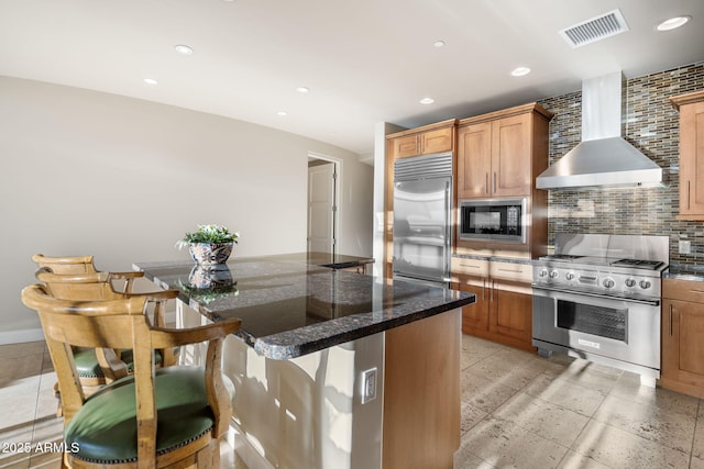 kitchen with a kitchen breakfast bar, backsplash, wall chimney range hood, and built in appliances