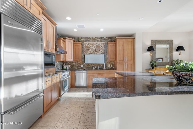 kitchen with wall chimney exhaust hood, tasteful backsplash, built in appliances, and dark stone countertops