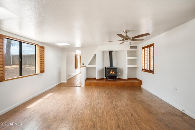 unfurnished living room featuring a wood stove, a ceiling fan, wood finished floors, and baseboards