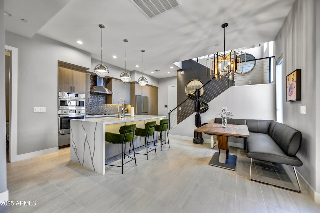 kitchen with sink, decorative light fixtures, a center island with sink, wall chimney range hood, and backsplash